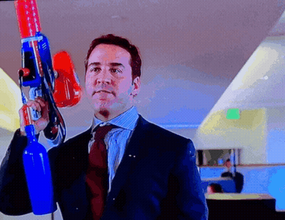 A Man In A Suit And Tie Holds A Large Toy Gun GIF