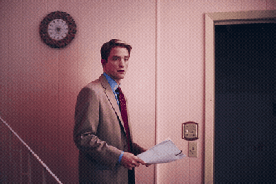 A Man In A Suit And Tie Holds A Piece Of Paper In Front Of A Pink Wall GIF