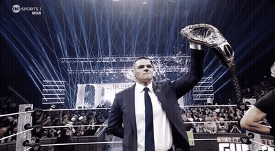 A Man In A Suit And Tie Holds Up A Wrestling Championship Belt In Front Of A Crowd GIF