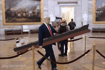 A Man In A Suit And Tie Is Carrying A Model Of A Ship That Says ' Donald Trump ' On It GIF