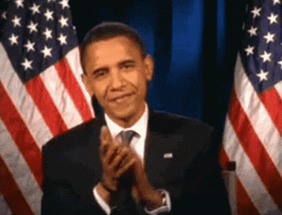 A Man In A Suit And Tie Is Clapping In Front Of American Flags . GIF