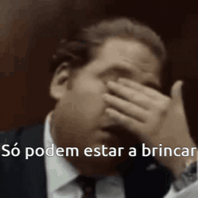 A Man In A Suit And Tie Is Covering His Face With His Hand And Says `` So Podem Estar A Brincar '' . GIF