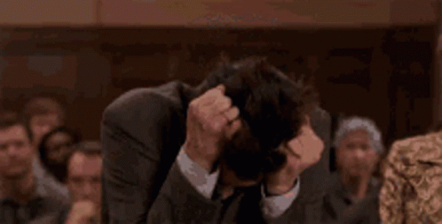 A Man In A Suit And Tie Is Covering His Face With His Hands In Front Of A Crowd Of People . GIF