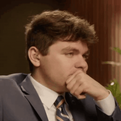 A Man In A Suit And Tie Is Covering His Mouth With His Hand While Sitting In A Chair . GIF