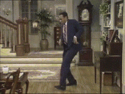 A Man In A Suit And Tie Is Dancing In A Living Room With A Clock In The Background GIF