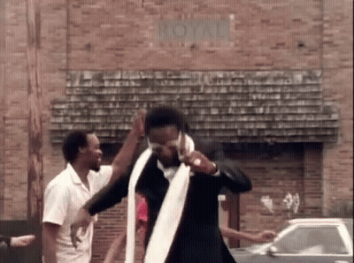 A Man In A Suit And Tie Is Dancing In Front Of A Brick Building With A Car In The Background . GIF