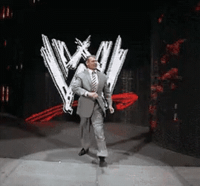 A Man In A Suit And Tie Is Dancing In Front Of A Wrestling Logo GIF