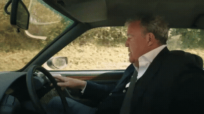 A Man In A Suit And Tie Is Driving A Car On A Dirt Road GIF