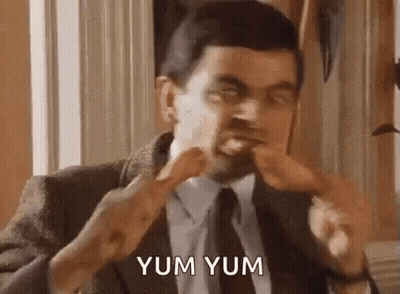 A Man In A Suit And Tie Is Eating A Chicken Leg And Saying Yum Yum . GIF