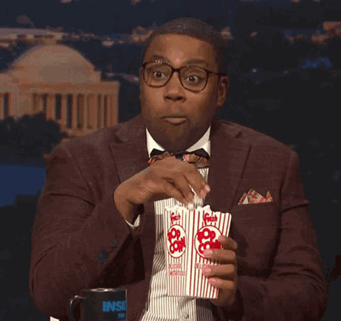 A Man In A Suit And Tie Is Eating Popcorn From A Container That Says Inside GIF