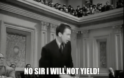 A Man In A Suit And Tie Is Giving A Speech In Front Of A Crowd Of People In A Courtroom . GIF