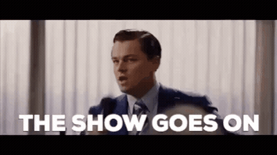 A Man In A Suit And Tie Is Giving A Speech In Front Of A Microphone And Saying The Show Goes On . GIF