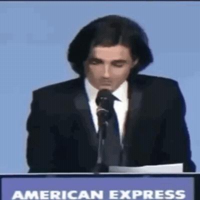A Man In A Suit And Tie Is Giving A Speech In Front Of An American Express Sign GIF