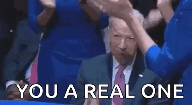 A Man In A Suit And Tie Is Giving A Thumbs Up While Sitting In A Crowd Of People . GIF