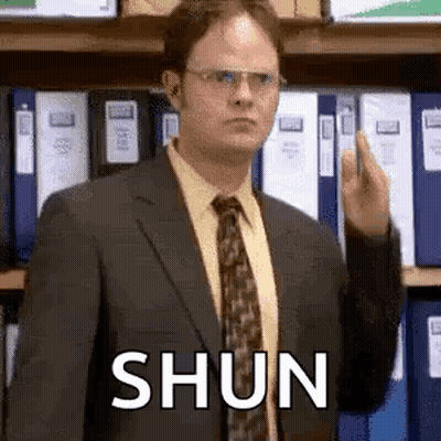 A Man In A Suit And Tie Is Giving The Middle Finger In Front Of A Shelf Of Folders . GIF