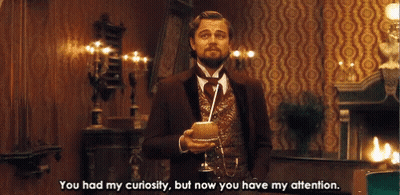 A Man In A Suit And Tie Is Holding A Glass With A Straw And Says You Had My Curiosity But Now You Have My Attention GIF