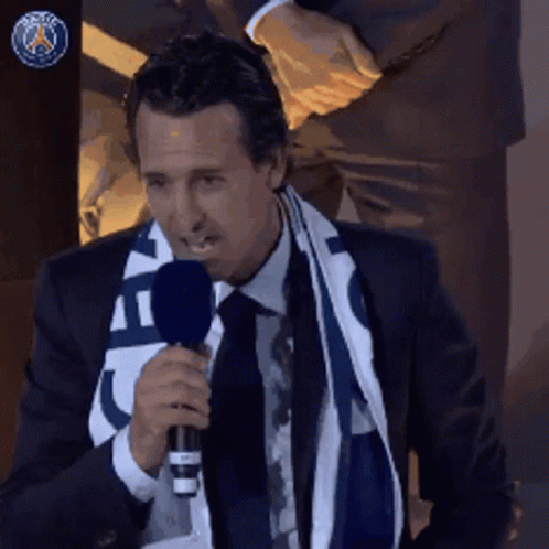 A Man In A Suit And Tie Is Holding A Microphone And Wearing A Scarf That Says Paris GIF
