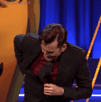 A Man In A Suit And Tie Is Holding A Microphone In Front Of A Blue Curtain That Says Itsscottiesstark On It GIF