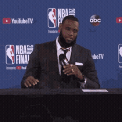 A Man In A Suit And Tie Is Holding A Microphone In Front Of A Nba Finals Banner GIF