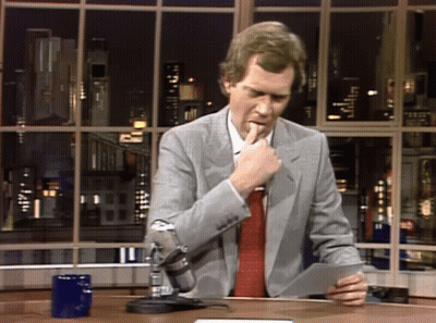 A Man In A Suit And Tie Is Holding A Piece Of Paper In Front Of A Window GIF