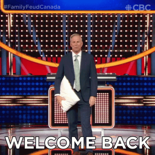 A Man In A Suit And Tie Is Holding A Pillow In Front Of A Stage That Says Welcome Back GIF