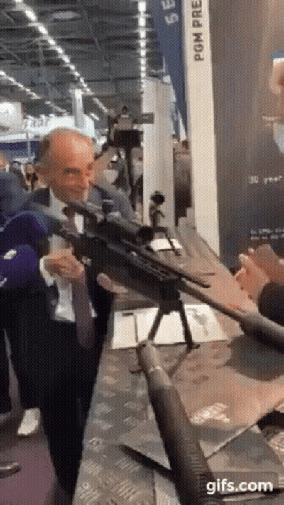 A Man In A Suit And Tie Is Holding A Rifle In Front Of A Camera . GIF
