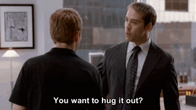 A Man In A Suit And Tie Is Hugging Another Man In A Black Shirt In An Office . GIF