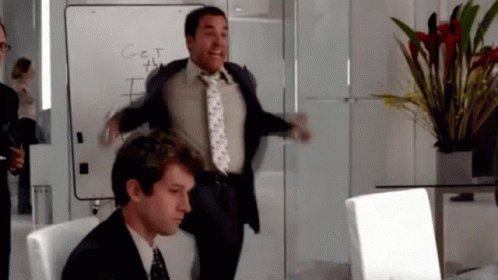 A Man In A Suit And Tie Is Jumping In The Air In Front Of A White Board That Says Get GIF