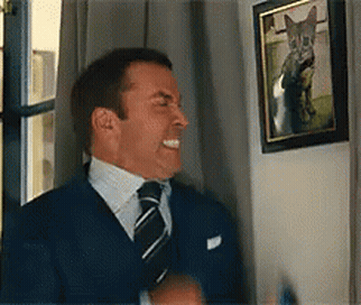 A Man In A Suit And Tie Is Laughing In Front Of A Picture Of A Cat On A Wall . GIF