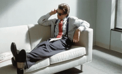 A Man In A Suit And Tie Is Laying On A White Couch With His Feet Up . GIF