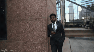 A Man In A Suit And Tie Is Leaning Against A Building While Holding A Cup Of Coffee GIF