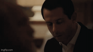 A Man In A Suit And Tie Is Looking At A Woman In A Room . GIF