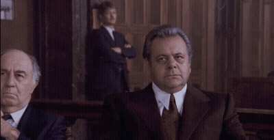 A Man In A Suit And Tie Is Looking At The Camera While Another Man Stands Behind Him . GIF