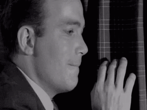 A Man In A Suit And Tie Is Peeking Out Of A Window With His Hand On The Curtain . GIF