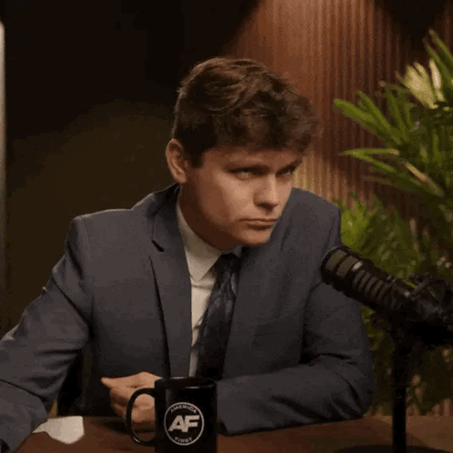 A Man In A Suit And Tie Is Pointing At A Mug That Says Af GIF