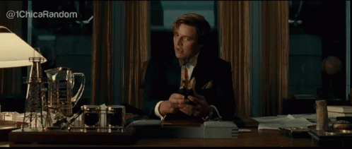 A Man In A Suit And Tie Is Sitting At A Desk And Looking At His Phone GIF