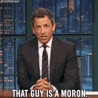 A Man In A Suit And Tie Is Sitting At A Desk And Says That Guy Is A Moron GIF