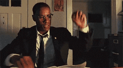 A Man In A Suit And Tie Is Sitting At A Desk Holding A Piece Of Paper . GIF