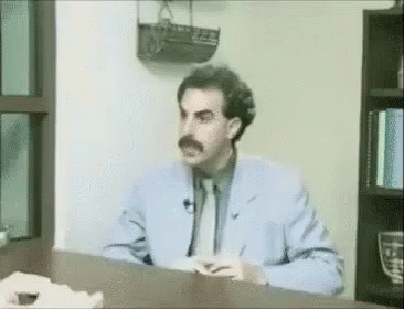 A Man In A Suit And Tie Is Sitting At A Desk In Front Of A Bookshelf . GIF