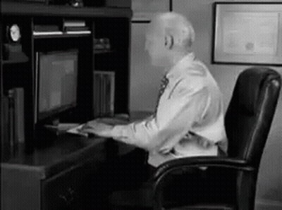 A Man In A Suit And Tie Is Sitting At A Desk In Front Of A Computer Monitor . GIF