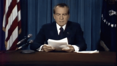 A Man In A Suit And Tie Is Sitting At A Desk In Front Of A Flag GIF