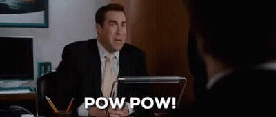 A Man In A Suit And Tie Is Sitting At A Desk In Front Of A Laptop Computer And Talking To Another Man . GIF
