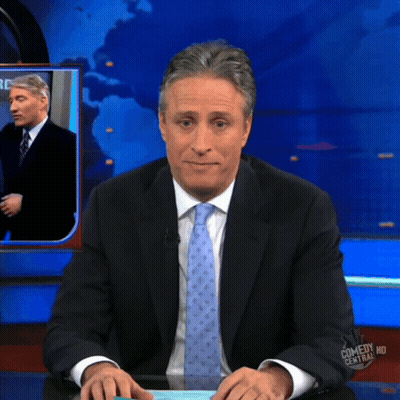 A Man In A Suit And Tie Is Sitting At A Desk In Front Of A Screen That Says Comedy Central Hd GIF