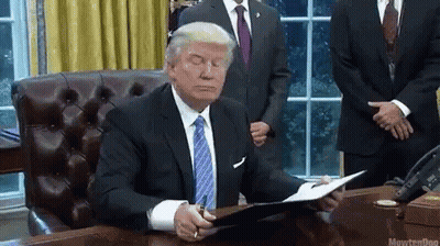 A Man In A Suit And Tie Is Sitting At A Desk Reading A Book . GIF