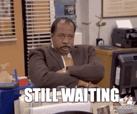 A Man In A Suit And Tie Is Sitting At A Desk With His Arms Crossed And The Words Still Waiting Below Him GIF
