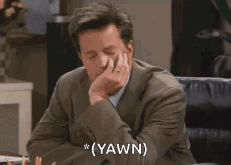 A Man In A Suit And Tie Is Sitting At A Desk With His Hand On His Chin And Says `` Yawn '' . GIF
