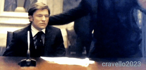 A Man In A Suit And Tie Is Sitting At A Desk With The Name Cravello2023 Written On The Bottom Right GIF