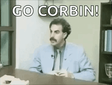 A Man In A Suit And Tie Is Sitting At A Desk With The Words `` Go Corbin '' Written On It . GIF