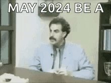 A Man In A Suit And Tie Is Sitting At A Desk With The Words `` May 2024 Be A '' Written On It . GIF