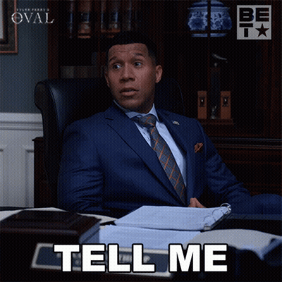 A Man In A Suit And Tie Is Sitting At A Desk With The Words Tell Me Below Him GIF